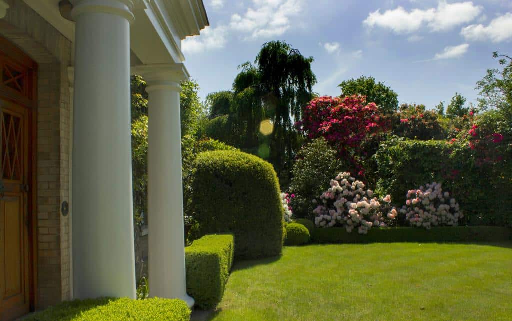 Richard Stone 1 Lush green lawn and neatly trimmed hedges surround a classical-style entrance, with vibrant flowering bushes and a clear blue sky.