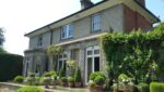 Richard Stone 2 Charming two-story house adorned with climbing plants, surrounded by neatly trimmed hedges and decorative potted plants in a sunny garden.