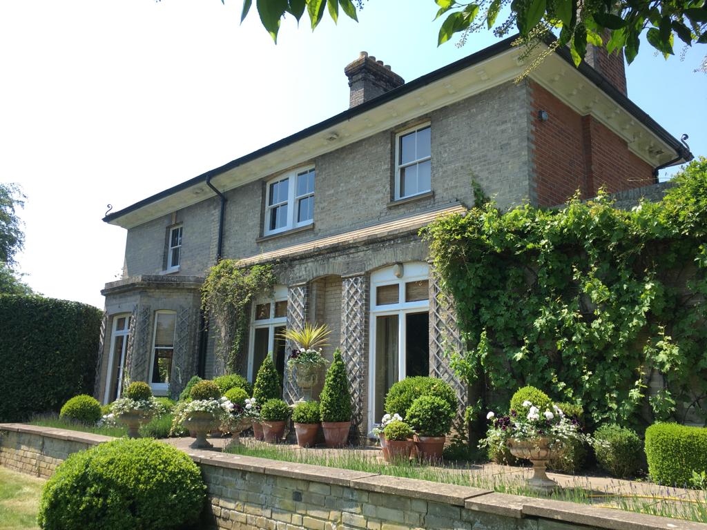 Richard Stone 2 Charming two-story house adorned with climbing plants, surrounded by neatly trimmed hedges and decorative potted plants in a sunny garden.