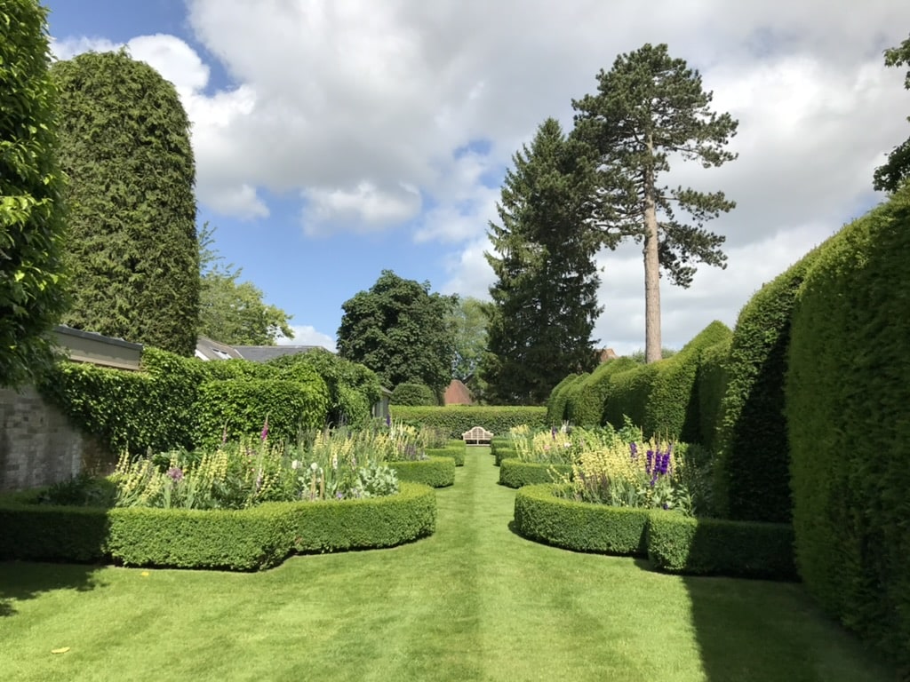 Richard Stone 4 Lush manicured garden with neatly trimmed hedges, vibrant flowers, and a tall tree under a partly cloudy sky.