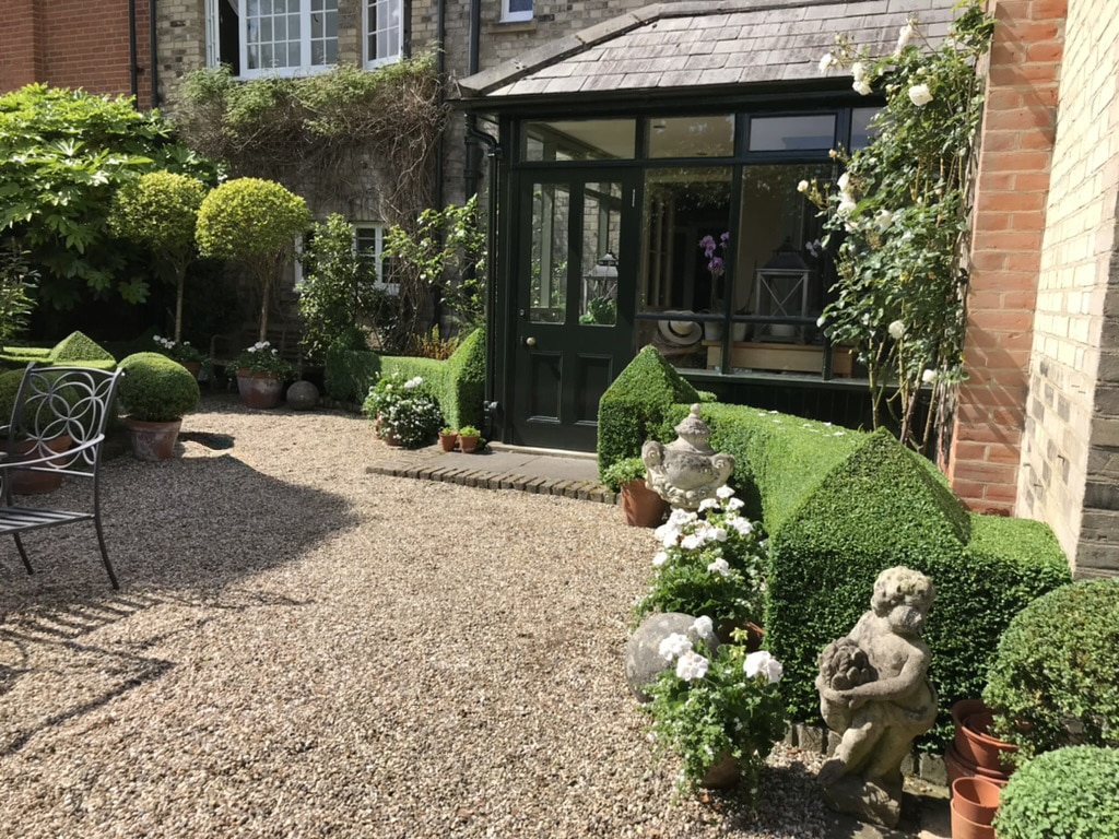 Richard Stone 9 Well-trimmed hedges, potted flowers, and a stone figure create an inviting garden space beside a green garden shed.