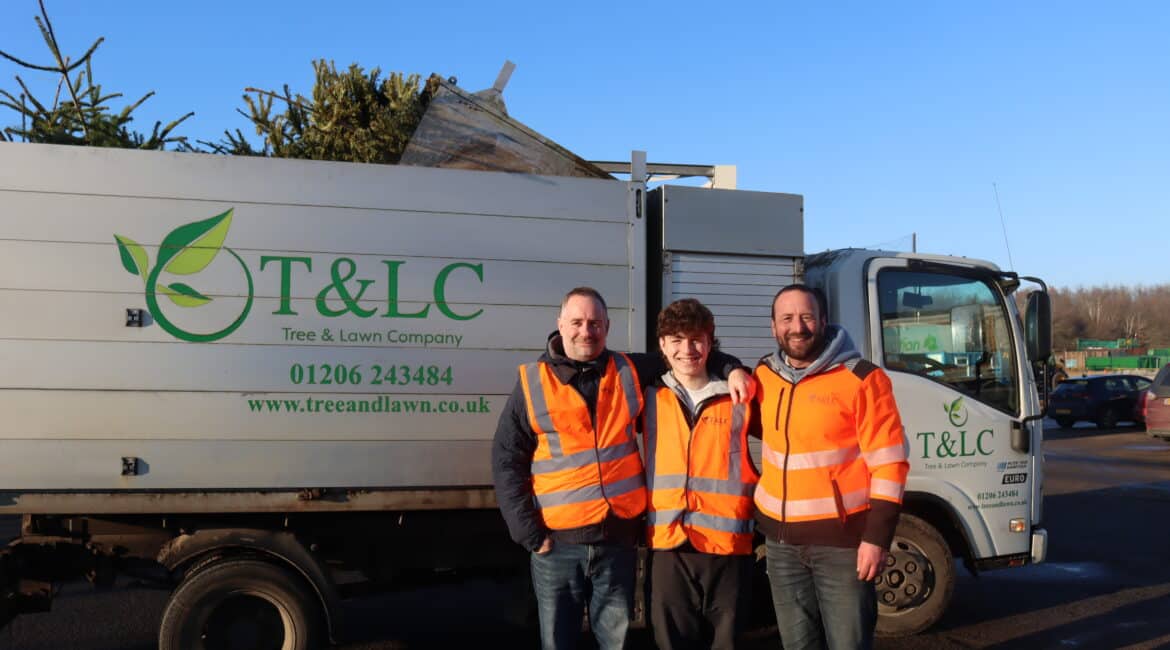 t l 2 Three workers in bright orange safety vests stand smiling in front of a truck with a logo for Tree & Lawn Company, loaded with Christmas trees.